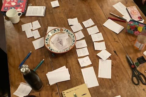 A picture of a table covered in index cards, pens, rulebooks, and other TTRPG things